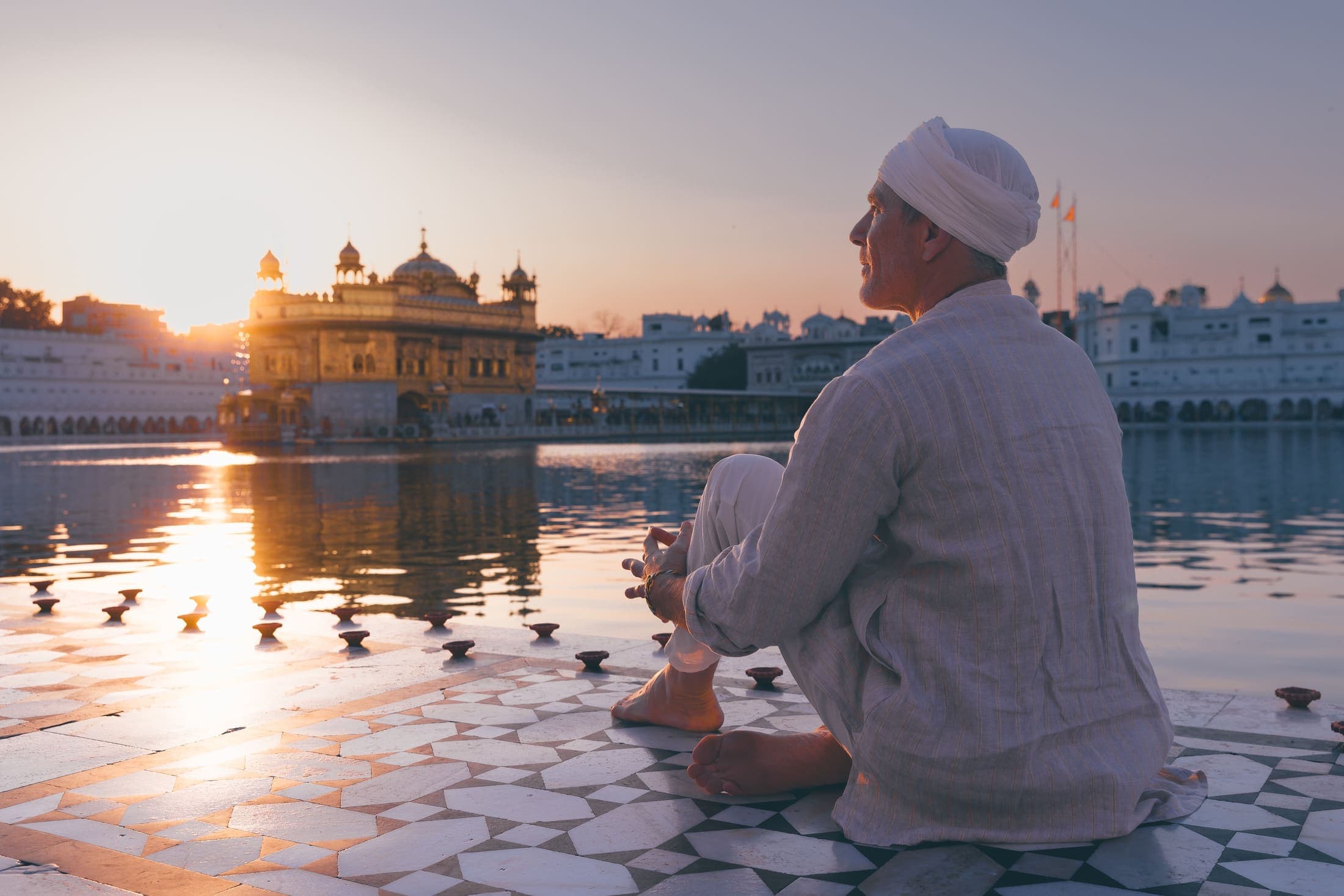 A man in a white turban seated in quiet reflection beside a temple reflecting pool at golden hour