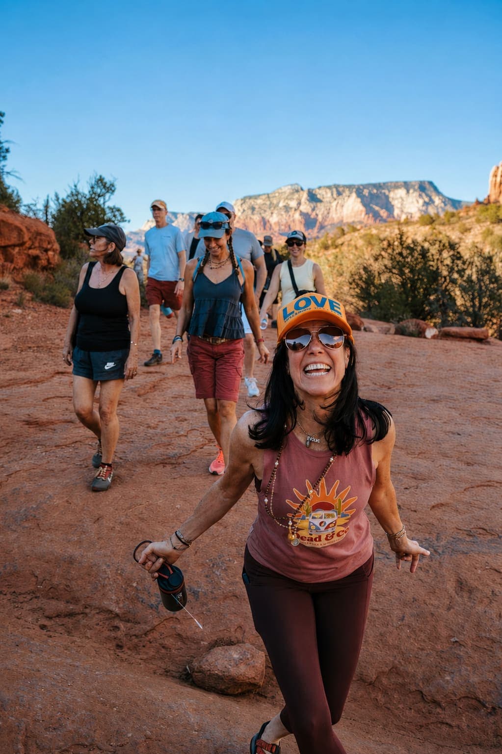 A joyful woman in an orange LOVE cap leads a group hiking together on a red rock trail
