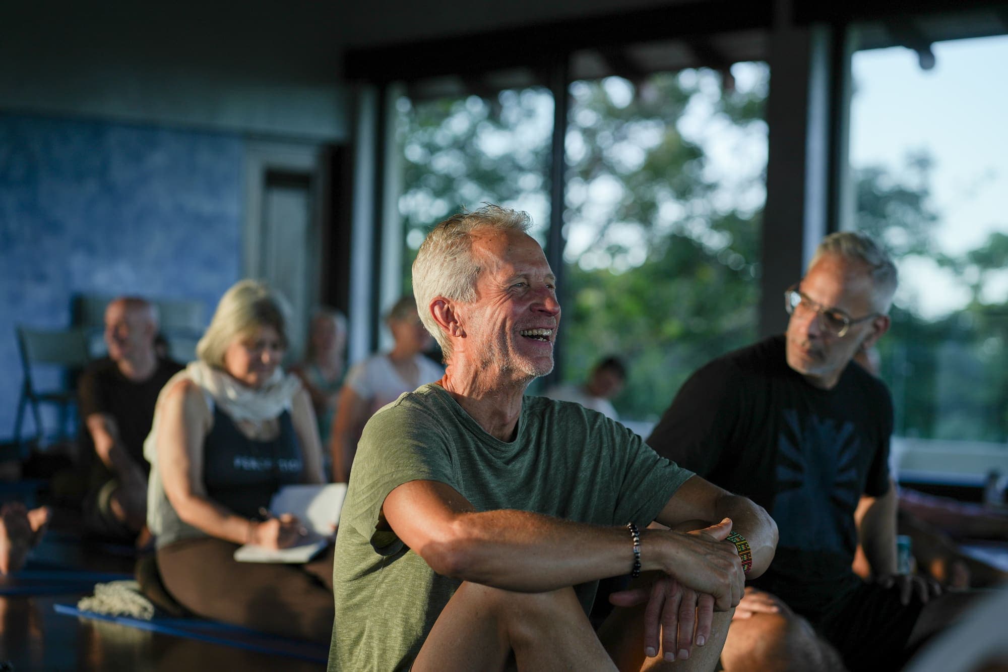 A silver-haired man smiling and looking off to the side during a retreat gathering, warm evening light