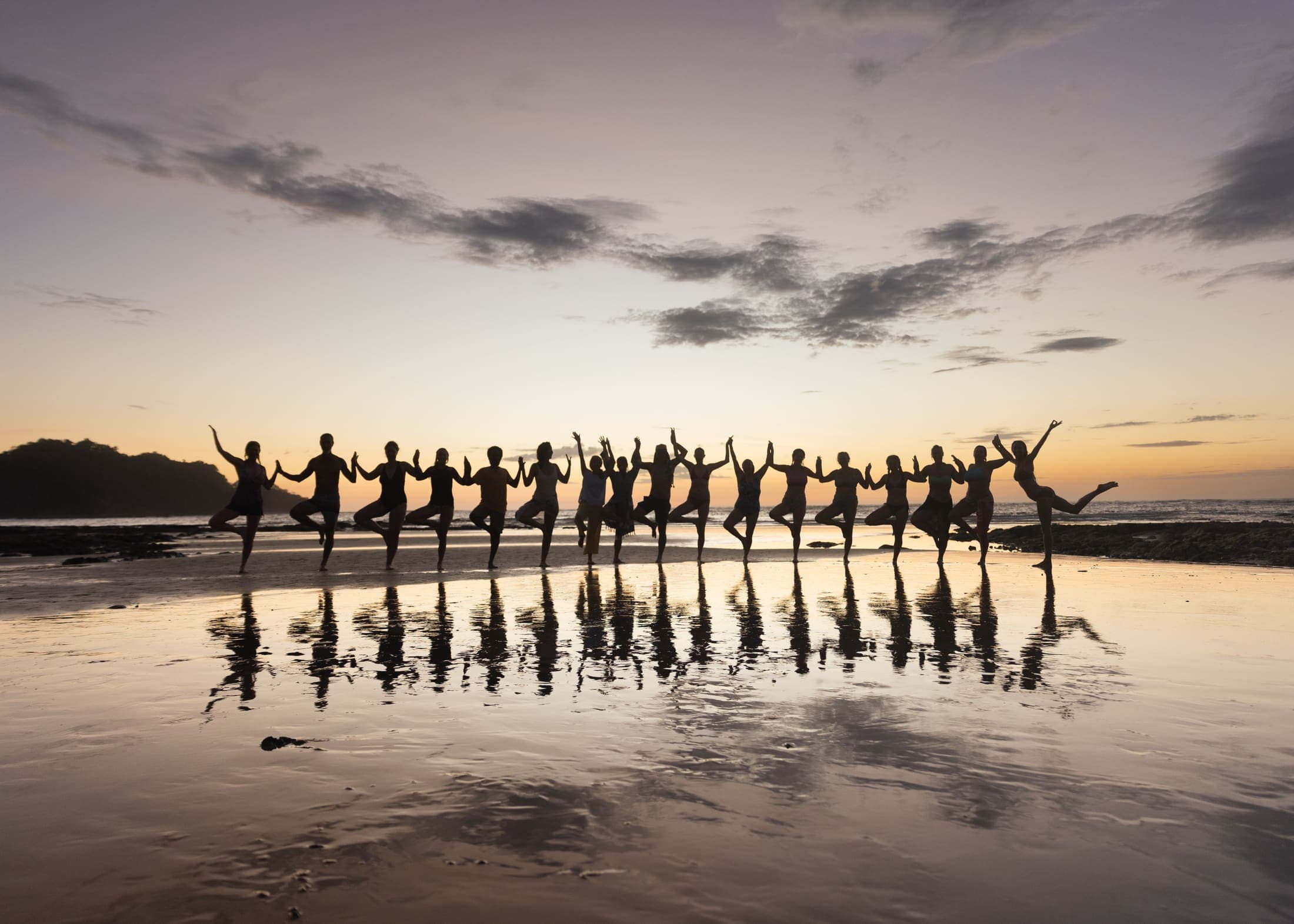 A large group holding hands in tree pose on a beach at sunset, silhouetted with a mirror-like reflection in the wet sand