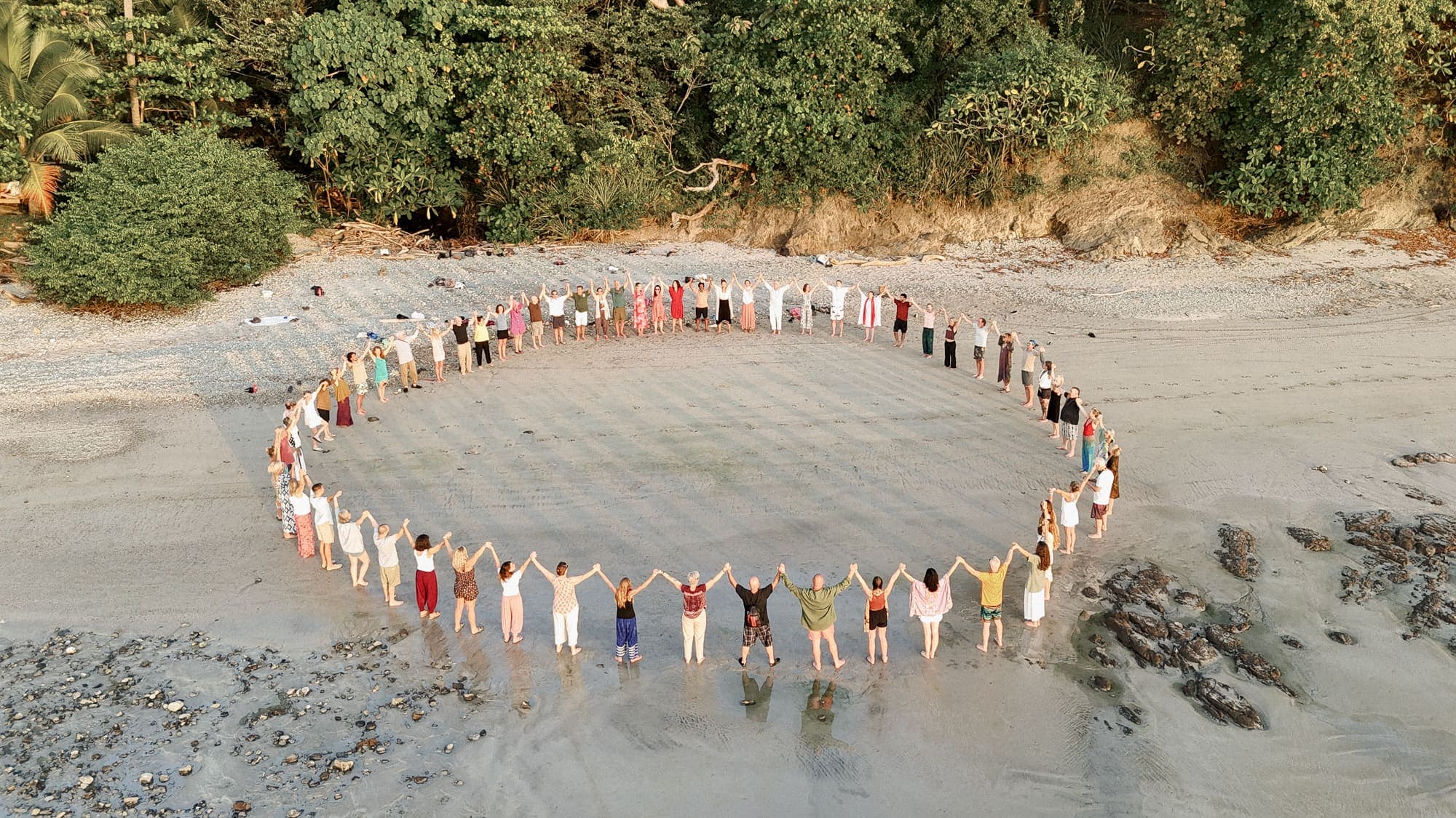 Aerial view of a large circle of people holding hands together on a beach at golden hour, symbolizing unity and shared purpose
