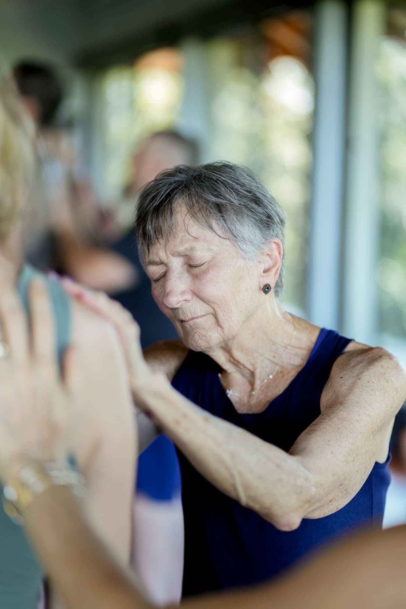An older woman with eyes closed, hand extended in a tender energy-healing gesture over someone else