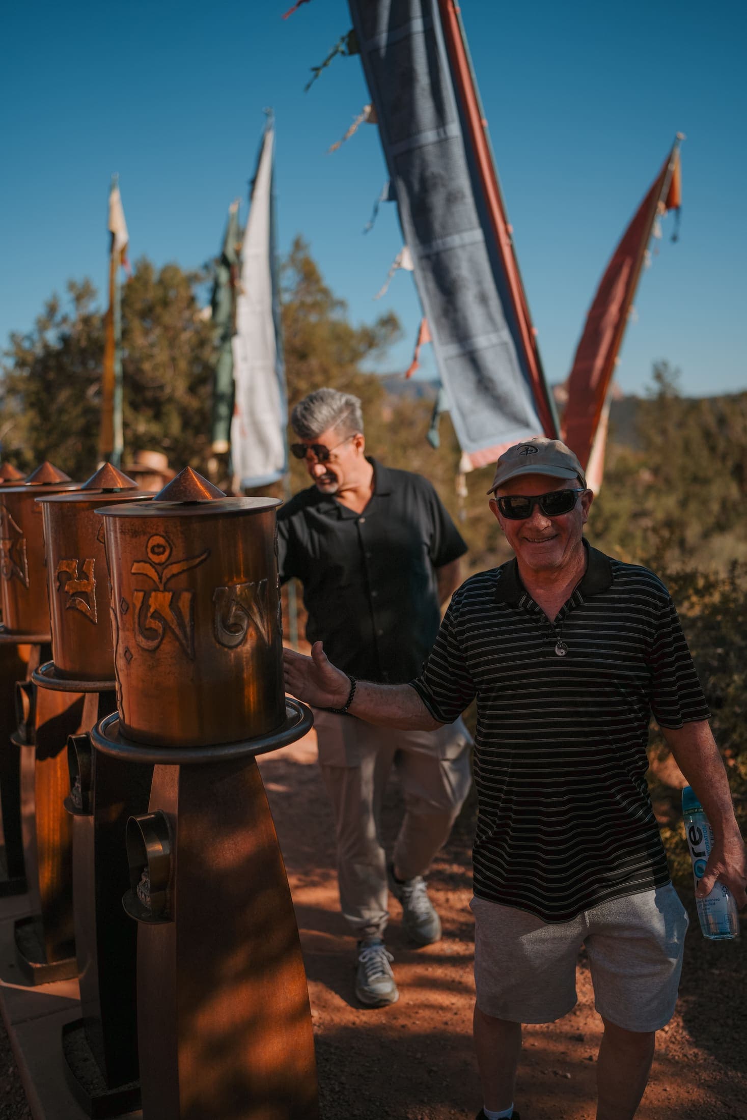 Two men laughing together beside copper prayer wheels and prayer flags in Sedona